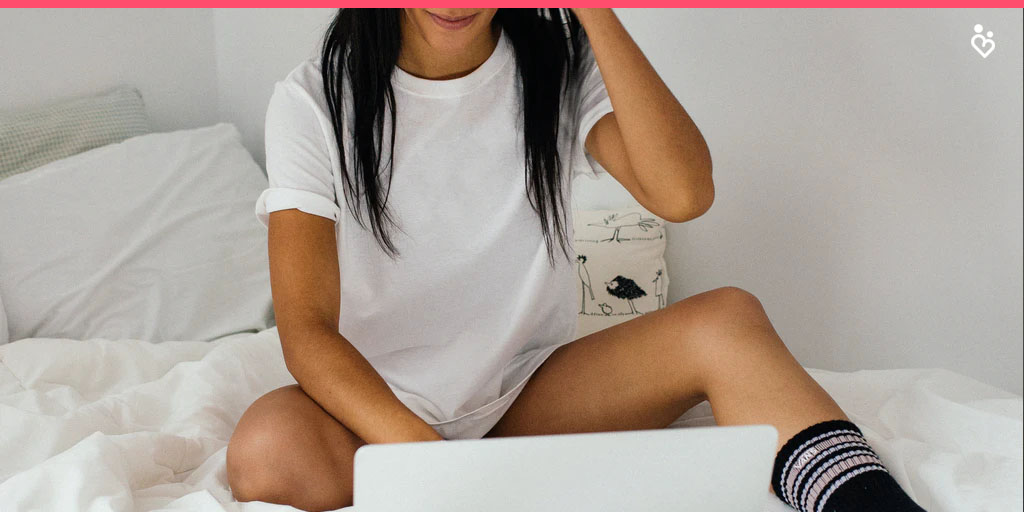 Chica con medias negras y franela blanca sentada en la cama chateando con su laptop